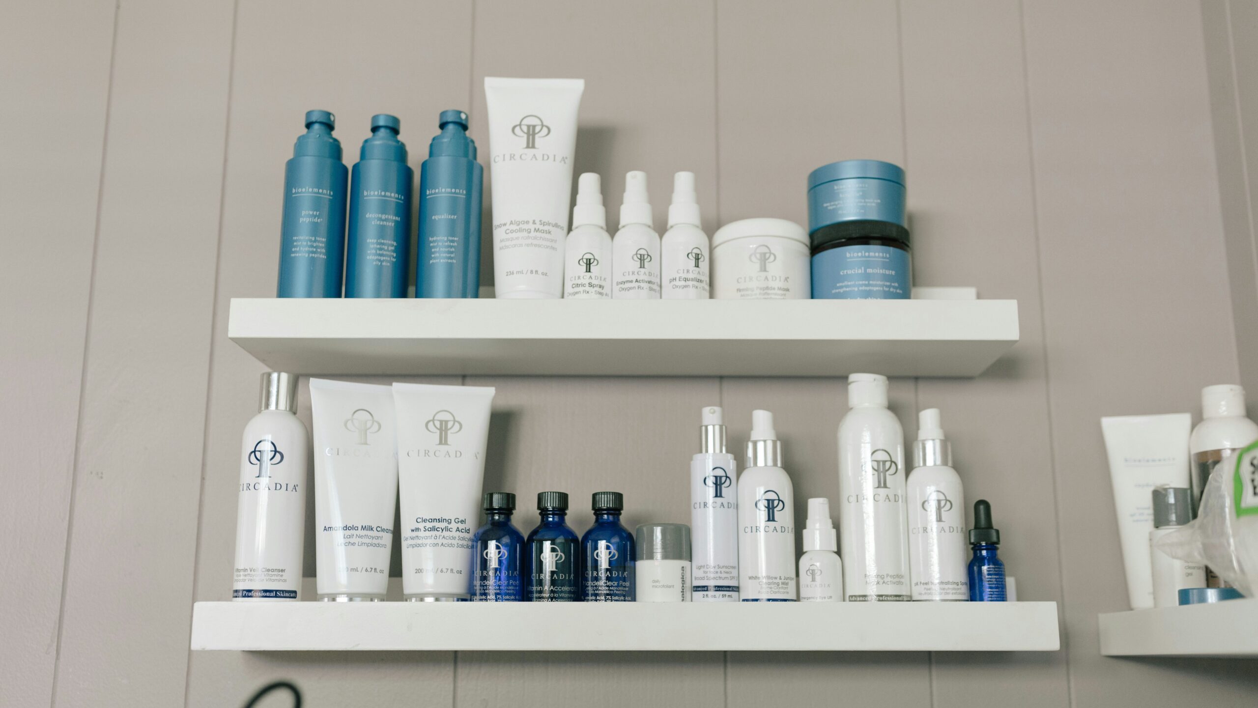 The image shows two white shelves mounted on a light gray wall, displaying a variety of skincare products. The products mainly belong to two brands: Circadia and Bioclinic. The Circadia products have white packaging, featuring their logo and text, while the Bioclinic products are packaged in blue containers. The items include bottles, tubes, jars, and sprays, indicating a range of skincare products like cleansers, masks, and moisturizers, all arranged neatly on the shelves.